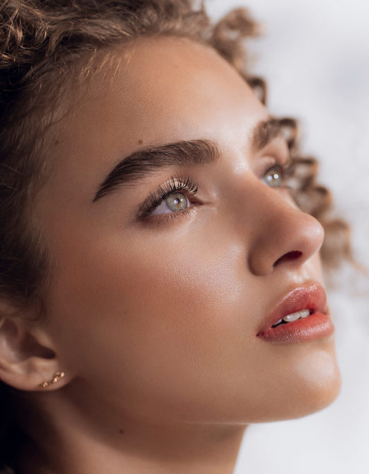 Eine Frau mit strahlender Haut, lockigem Haar, kräftigen Augenbrauen blickt nach oben und präsentiert Wirkung der Brow Refresh Collection von RevitaLash® Cosmetics – RevitaBrow Advanced Spring 2025.