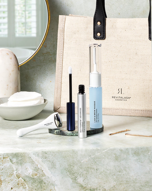 Die Ultimate Brow Collection – RevitaBrow® Advanced Summer 2024 von RevitaLash® Cosmetics steht auf einer Badezimmerablage, im Hintergrund eine beige Tasche und ein runder Spiegel.