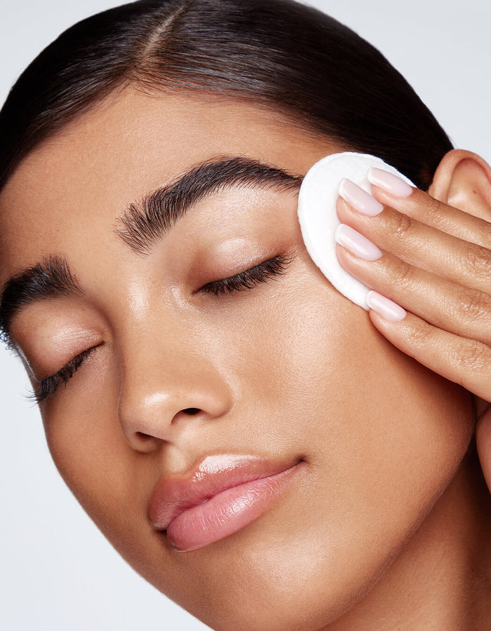 Eine Frau mit makelloser Haut wischt sich sanft mit einem Wattepad mit Micellar Water Lash Wash über das Gesicht, die Augen sind geschlossen.
