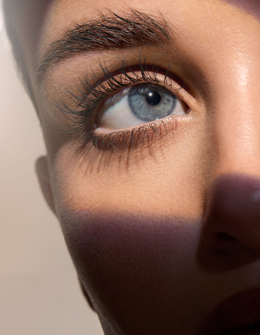 Close-up eines blauen Auges mit betonten, langen Wimpern und gepflegten Brauen, dramatisch ins Licht gesetzt durch Schatteneffekte.