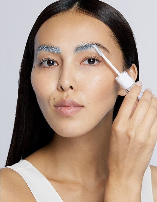 Eine Frau trägt vor einem schlichten Hintergrund mit einem Bürstchen die RevitaLash® Cosmetics Lash & Brow Masque auf ihre Augenbrauen auf.