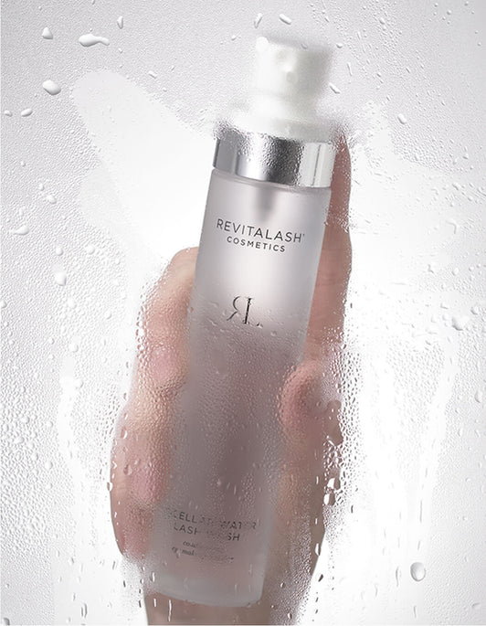 Eine Hand hält eine Flasche RevitaLash® Cosmetics Micellar Water Lash Wash hinter einer nebligen, nassen Glasoberfläche.