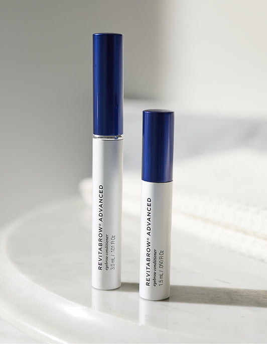 Zwei RevitaBrow® Advanced Eyebrow Conditioner-Tuben von RevitaLash® Cosmetics mit blauen Kappen werden in sanftem Licht auf eine weiße Oberfläche gestellt.