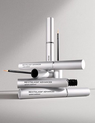 Mehrere silberne Tuben RevitaLash Advanced Eyelash Conditioner übereinander gestapelt auf einer grauen Oberfläche.