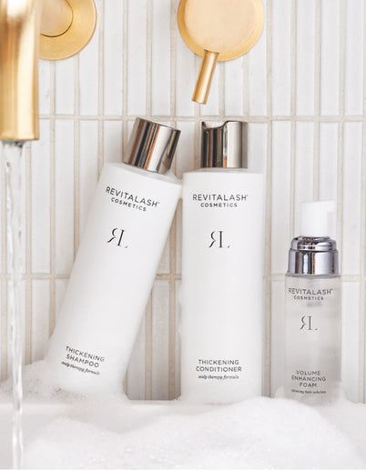 Drei Haarprodukte von RevitaLash Cosmetics auf einer weiß gefliesten Oberfläche mit Schaum und goldenen Wasserhähnen in der Nähe.
