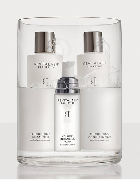 Drei Haarpflegeprodukte von RevitaLash® Cosmetics in einem durchsichtigen , mit Wasser gefülltem, Glasbehälter: Shampoo, Conditioner und Volume Foam.