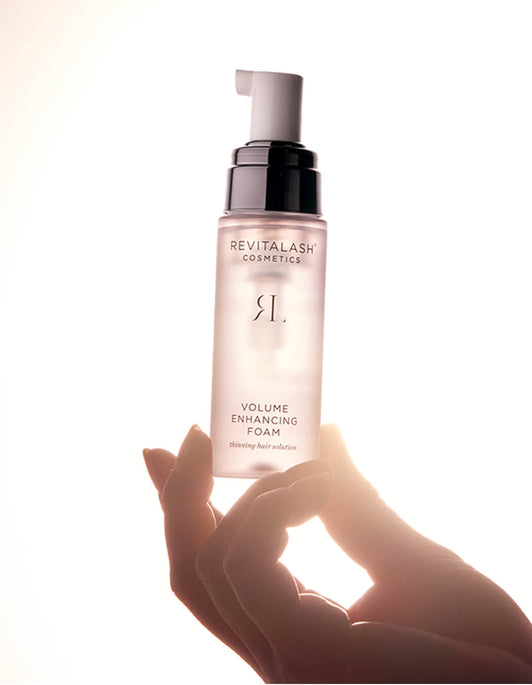 Eine Hand hält eine Flasche RevitaLash® Cosmetics Volume Enhancing Foam vor einem hellen, hinterleuchteten Hintergrund.