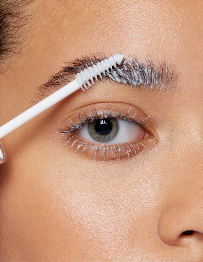 Eine Nahaufnahme einer Person, die mit einer weißen Spiralbürste Lash & Brow Masque auf ihre Augenbraue und Wimpern aufträgt.