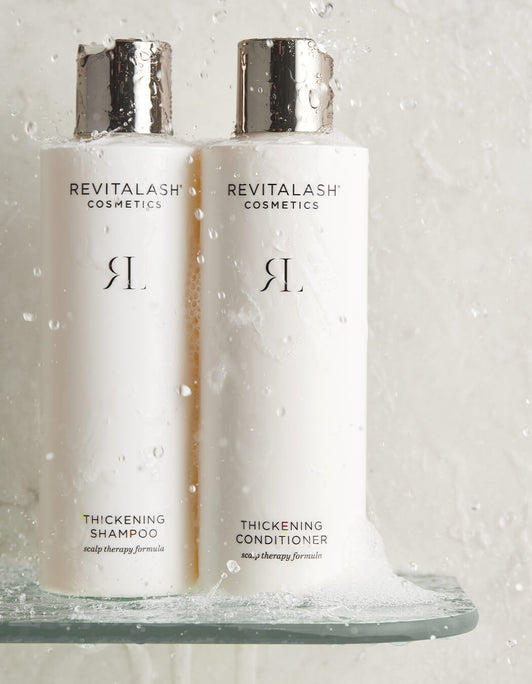 Zwei weiße Flaschen RevitaLash® Cosmetics Thickening Shampoo und Conditioner mit silbernen Deckeln stehen, bedeckt mit Schaum, auf einem nassen Duschregal.