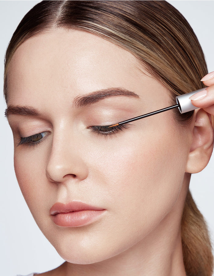 Eine Frau trägt mit einem kleinen Pinselapplikator RevitaLash Advanced Eyelash Serum auf ihre oberen Wimpern auf.