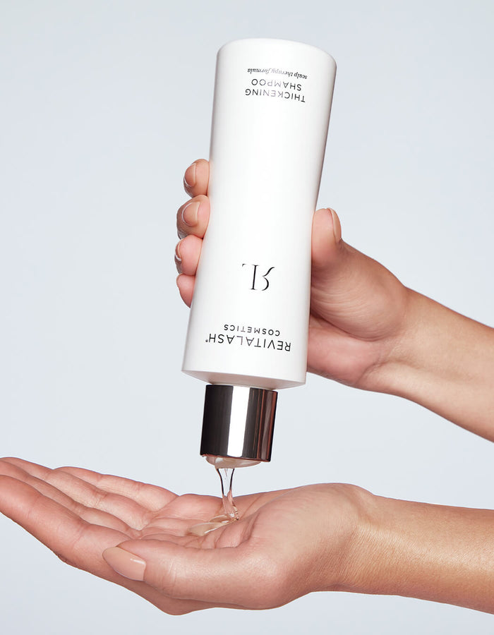 Hand gießt vor hellem Hintergrund klares Thickening Shampoo aus einer weißen RevitaLash-Flasche in eine offene Handfläche.