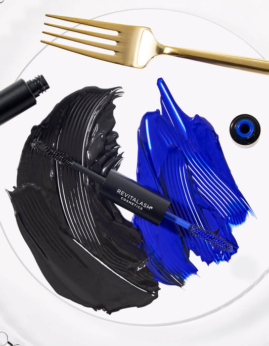 Auf weißem Teller liegt das RevitaLash Double-Ended Volume Set mit Primer und Mascara. Neben goldener Gabel sind schwarze und blaue Farbstriche aufgetragen, die die beiden Produktfarben zeigen.