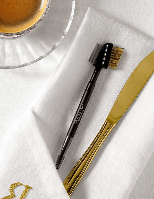 RevitaLash Lash & Brow Styler mit Bürstchen und Kamm liegt auf weißer Stoffserviette neben goldenem Besteck, oben im Bild eine Tasse Kaffee.