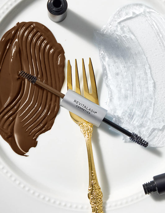 RevitaLash Double-Ended Hi-Def Brow Gel liegt auf einem weißen Teller mit goldener Gabel. Links brauner Farbstrich für Soft Brown, rechts transparenter Farbstrich für Clear.