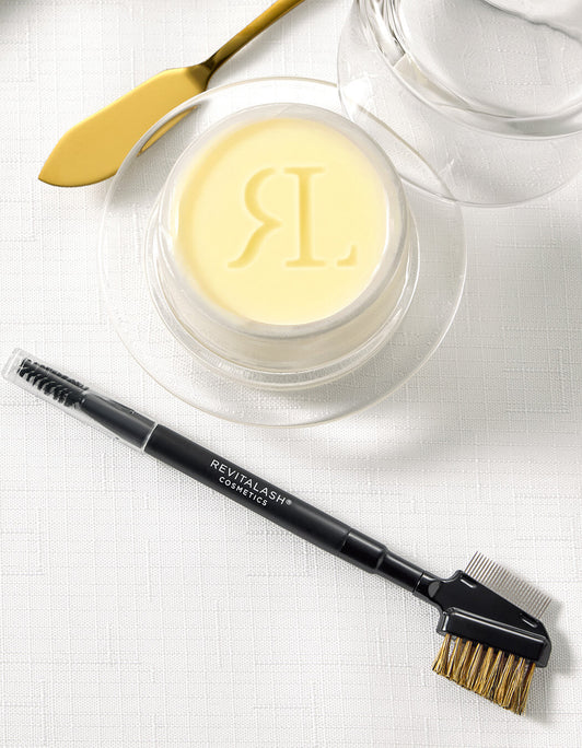 RevitaLash Lash & Brow Styler mit Bürstchen und Kamm liegt auf weißer Tischdecke neben einem Glas mit Butter und goldenem Messer.