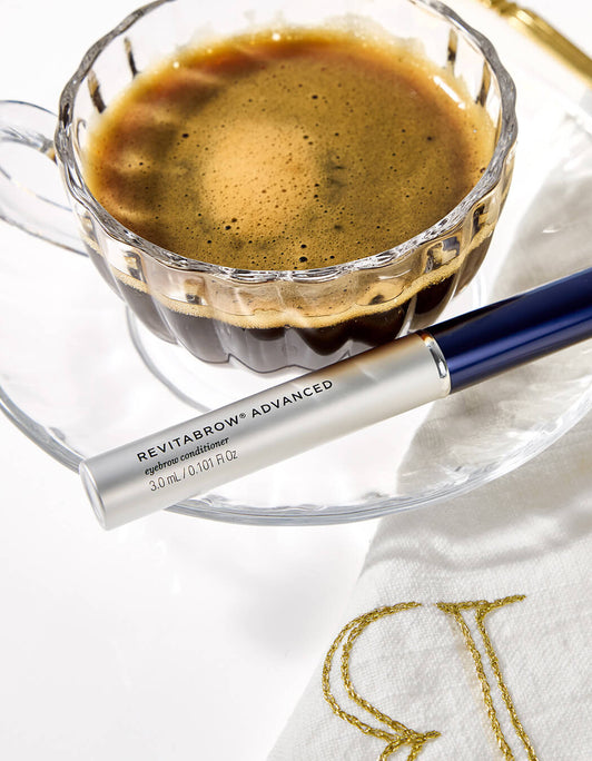 Eine Tube RevitaBrow® Advanced Eyebrow Conditioner liegt auf einer weißen Serviette mit goldener Stickerei neben einer Glas-Tasse Kaffee.
