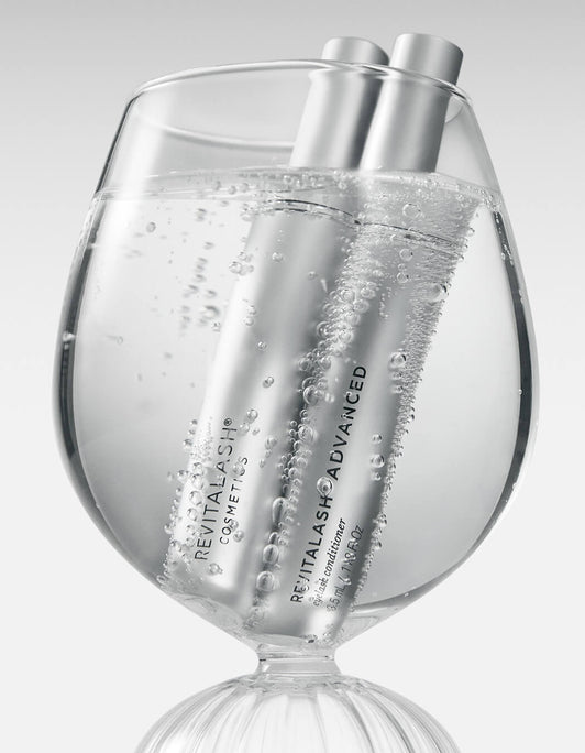 Zwei silberne Tuben RevitaLash® Advanced Eyelash Conditioner (3,5 ml) stehen in einem Glas mit sprudelndem Wasser, Bläschen steigen auf.