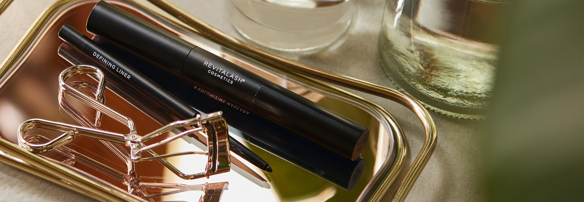 Eine goldene Wimpernzange und schwarze Make-up-Produkte auf einem reflektierenden Tablett neben Glasbehältern.
