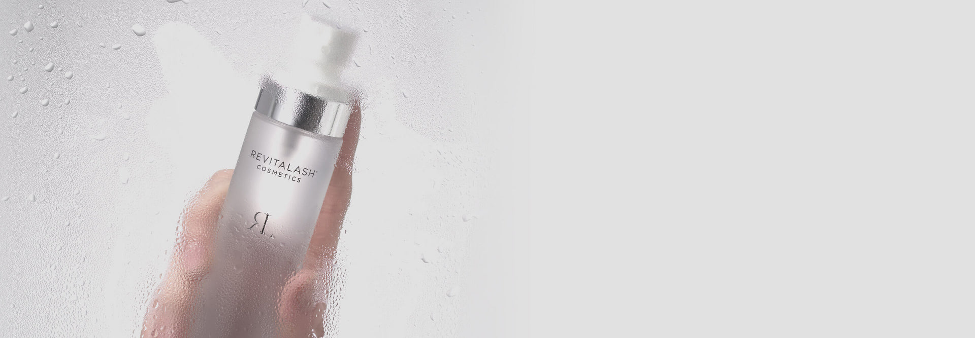 Hand hält eine Flasche Revitalash Cosmetics Micellar Water Lash Wash hinter einer nassen Glasoberfläche.