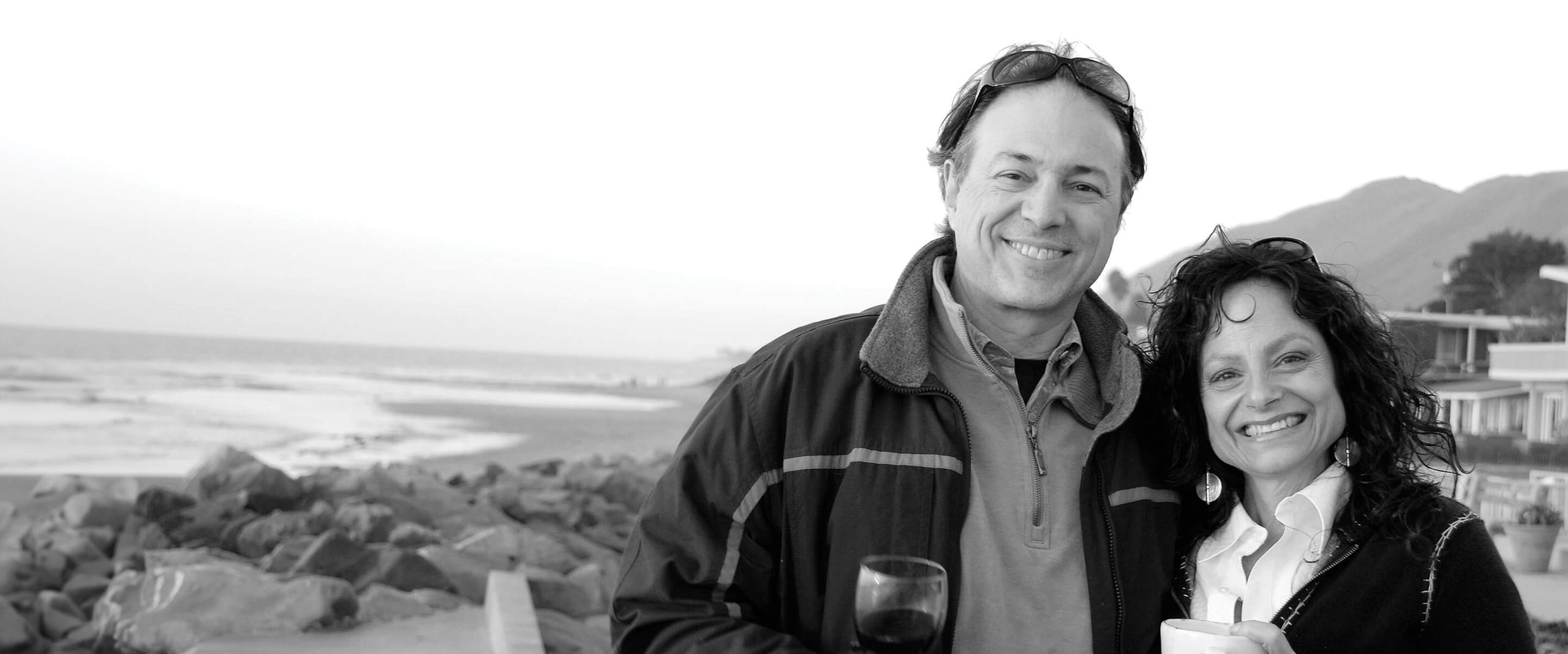 Schwarz-weiß Foto der lächelnden Gründer Michael und Gayle Brinkenhoff an einem felsigen Strand mit Bergen und Häusern im Hintergrund.