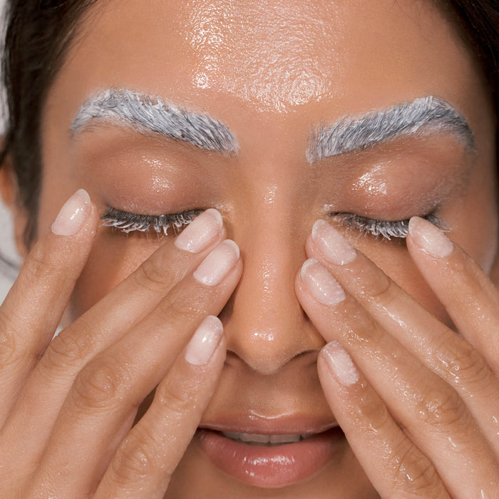 Eine Frau mit geschlossenen Augen trägt Reinigungsmittel auf ihr Gesicht auf, ihre Augenbrauen und Wimpern sind mit weißer Creme bedeckt.