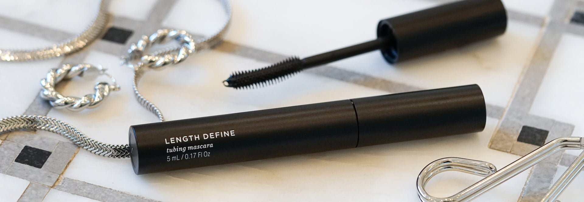 Schwarze Tube Length Define Tubing Mascara mit geschwungenem Bürstchen, Silberschmuck und Wimpernzange auf einer gefliesten Oberfläche.