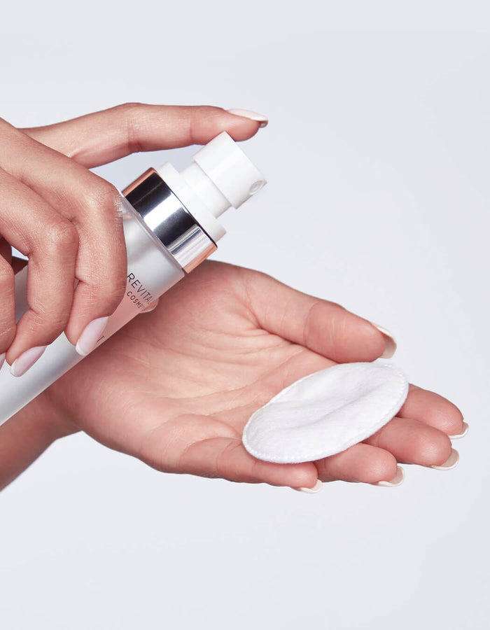 Vor einem hellen Hintergrund sprüht eine Hand ein Hautpflegeprodukt auf ein rundes Wattepad, das sie in der anderen Hand hält.