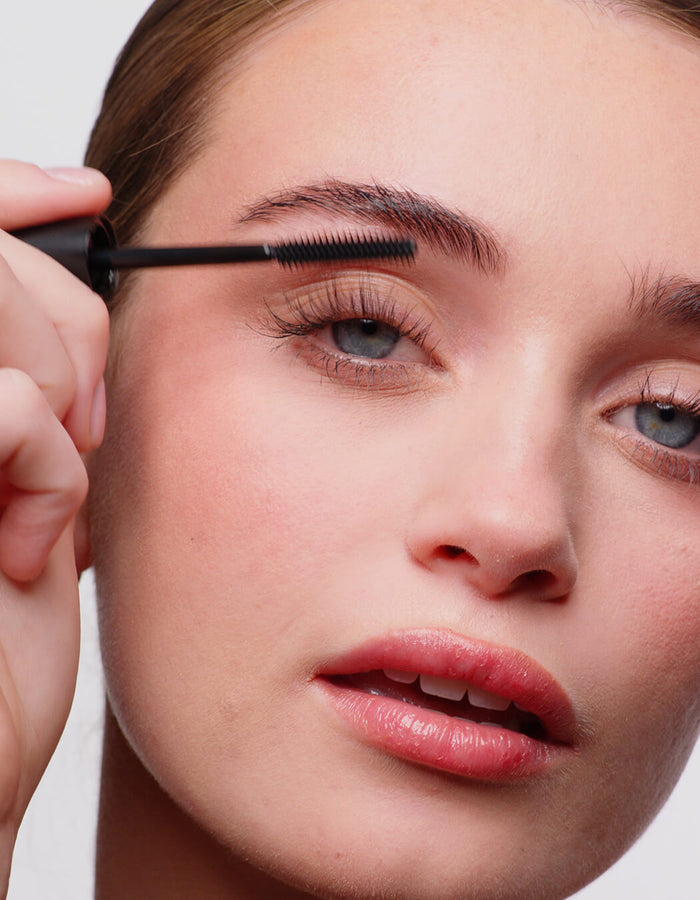 Frau trägt Mascara auf ihre Wimpern auf, Nahaufnahme ihres Gesichts mit natürlichem Make-up und rosa Lippen.