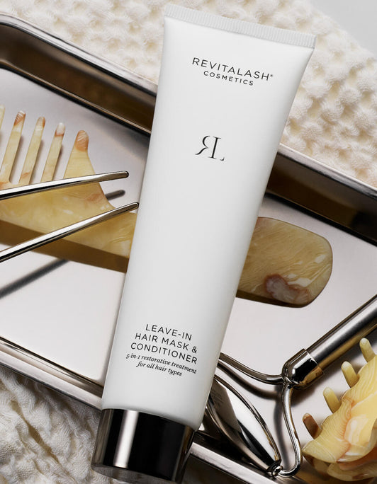 Eine Tube RevitaLash Germany Leave-In Hair Mask & Conditioner wird auf einem Metalltablett neben Haarspangen und Accessoires präsentiert.