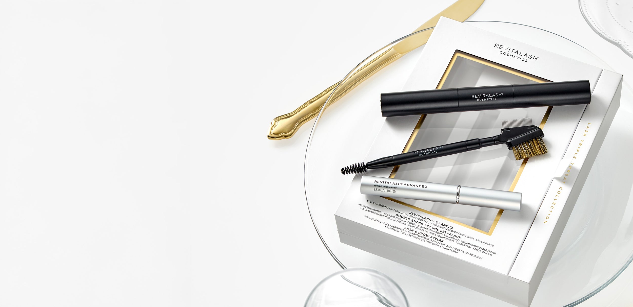 Das Lash Triple Threat Collection Set von RevitaLash® Cosmetics liegt auf einem weißen Teller mit goldfarbenem Besteck. Das Set enthält RevitaLash® Advanced Serum, die Double-Ended Volume Mascara und den Lash & Brow Styler.