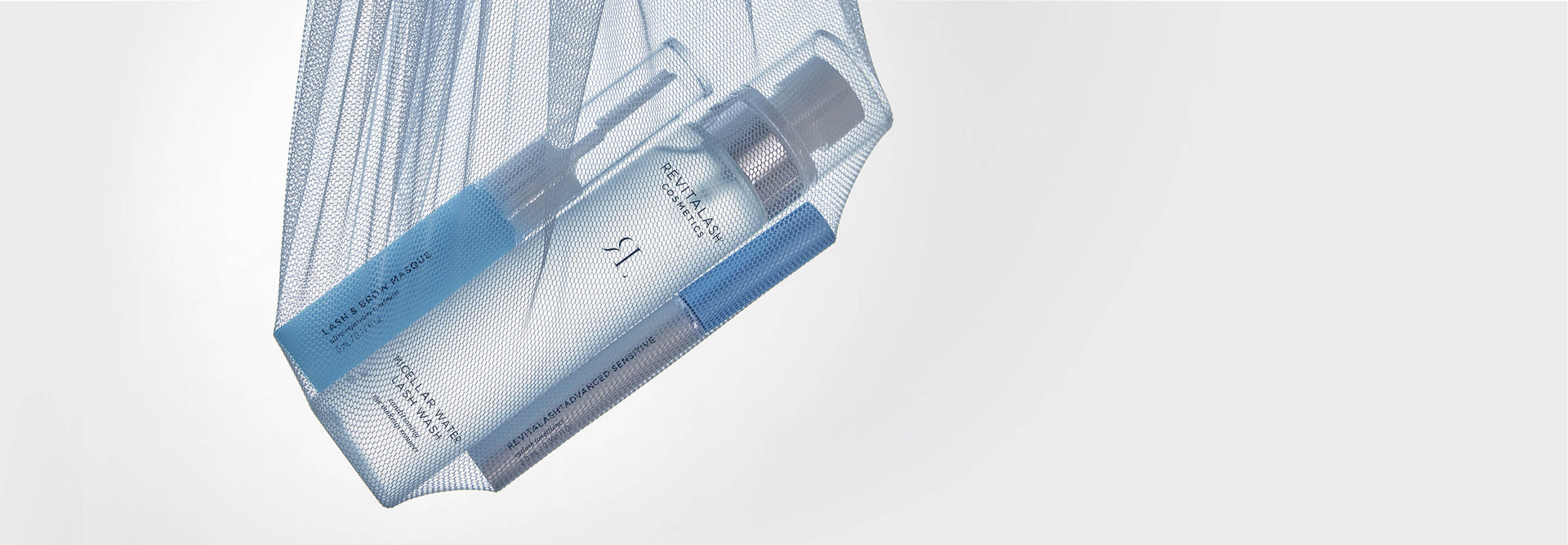Drei Kosmetikprodukte von RevitaLash® Cosmetics hängen in einem blauen Netzbeutel vor hellgrauem Hintergrund.