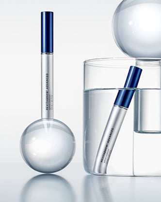 Zwei silber-blaue Tuben RevitaBrow® Advanced Eyebrow Conditioner mit Glaskugeln und einem Glas Wasser auf hellem, minimalistischem Hintergrund.