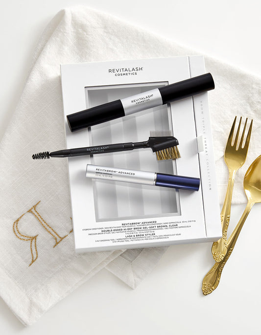 RevitaBrow Geschenkset mit Conditioner, Brow Gel und Styling-Tool, dekorativ inszeniert auf weißem Stoff mit goldenen Besteckakzenten.
