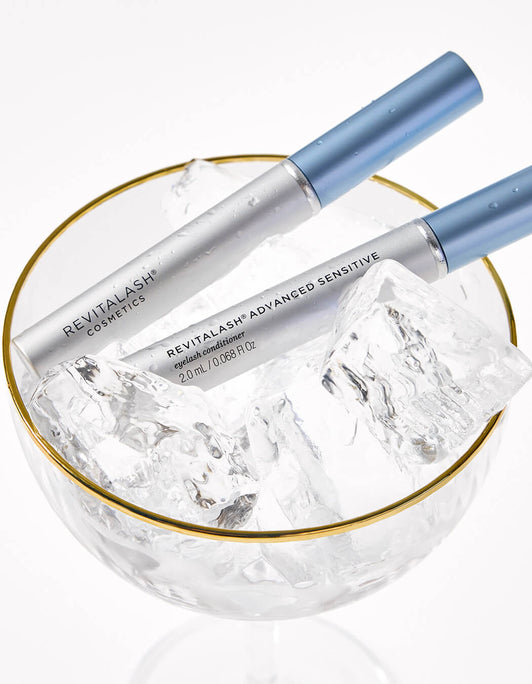 Zwei silber-blaue Tuben RevitaLash® Advanced Sensitive Eyelash Conditioner liegen in einem Glas mit Eiswürfeln und Goldrand.