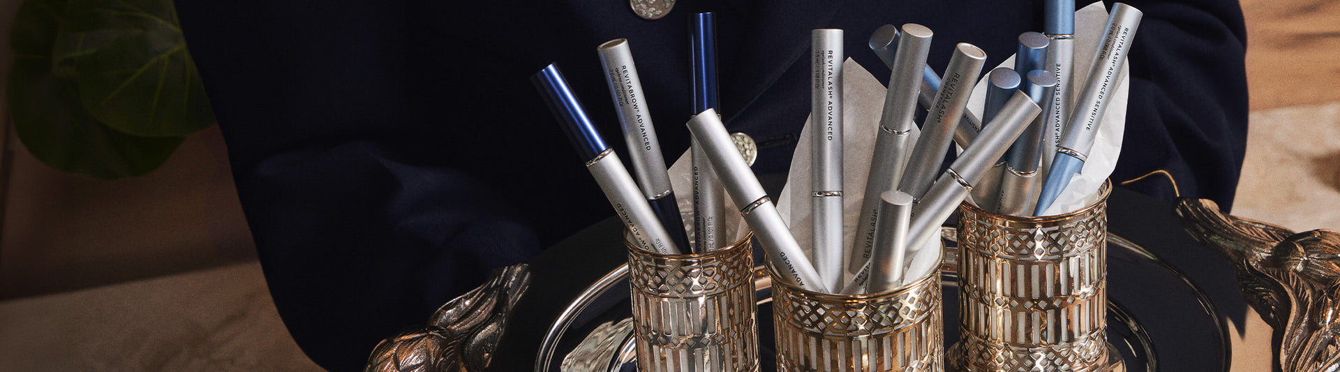Mehrere silberne RevitaLash® Advanced Conditioner präsentiert in dekorativen Metallbechern auf einem Tablett, elegant arrangiert in warmem Licht.