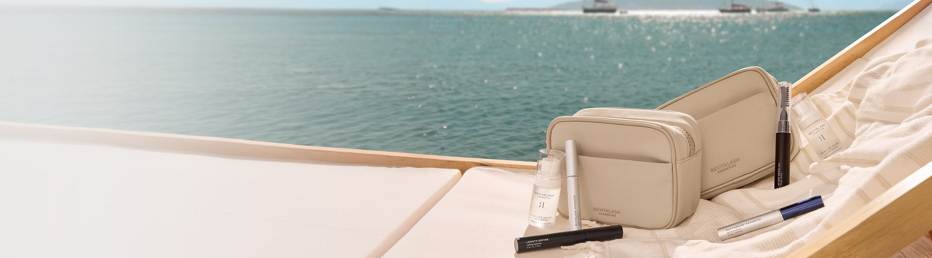 Beige Kosmetiktaschen und Schönheitsprodukte von RevitaLash auf einer beigen Sonnenliege mit Blick auf das ruhige blaue Meer an einem sonnigen Tag.